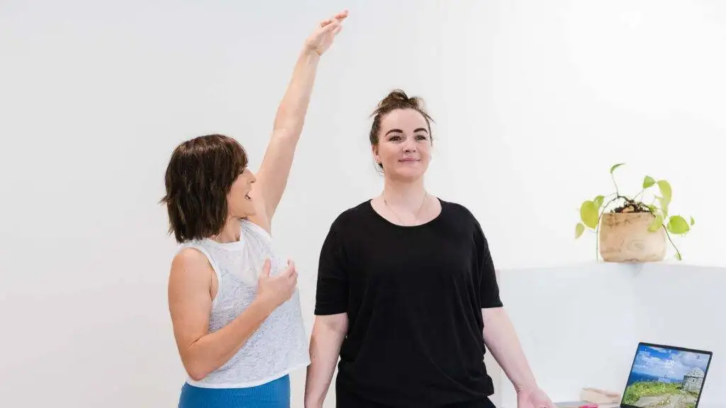 Two woman stretching next to a while wall in an office